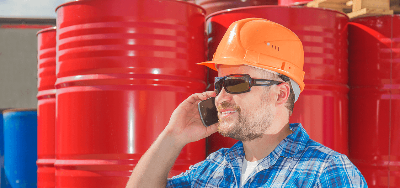 Man with sunglasses and an orange safety helmet talking on the phone in front of several red barrels.