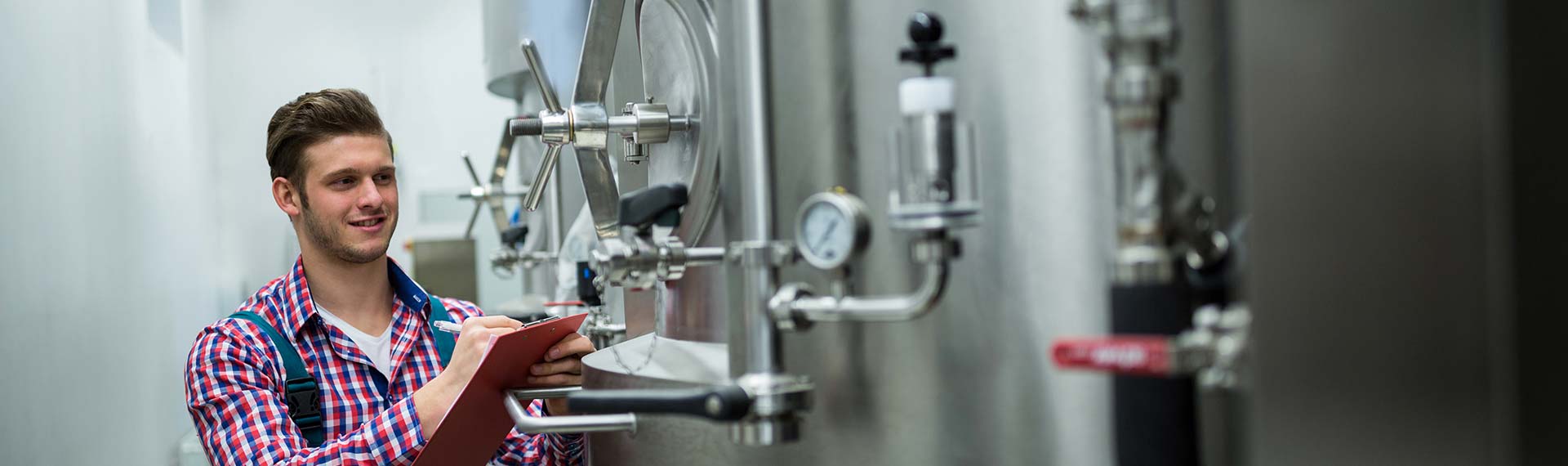 Young man taking notes in front of large metal containers in the food and beverage industry.